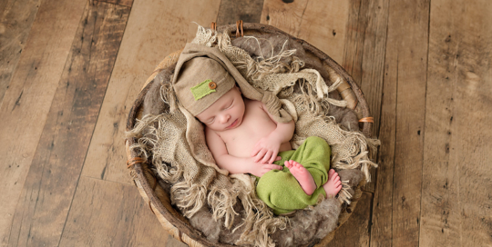 baby boy in green pants and sleepy hat in burlap and twig bucket on brown wood floor in kingston baby studio