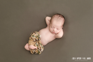 newborn baby boy on back with hands behind head in camo military pants in kingston ontario photography studio