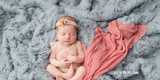 newborn baby girl in pink romper with pink flower headband with pink wrap on grey flokati rug in kingston ontario baby studio