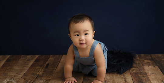 6 month asian baby boy in blue romper on wood floor with blue backdrop by kingston baby photographer