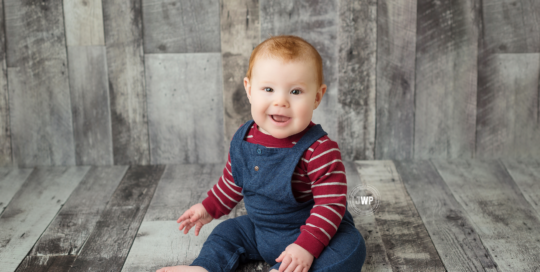 baby boy in blue overalls with red striped shirt on grey wood by kingston newborn photographer