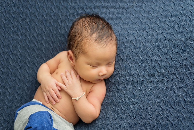 newborn baby boy close up laying on back on blue blanket with blue and grey pants and bracelet by Belleville Newborn Photographer