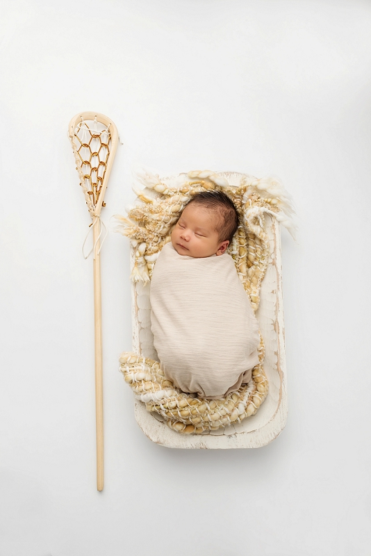 First Nations newborn baby in cream wrap on tan blanket on white wood trenchbowl with lacrosse stick by Belleville Newborn Photographer