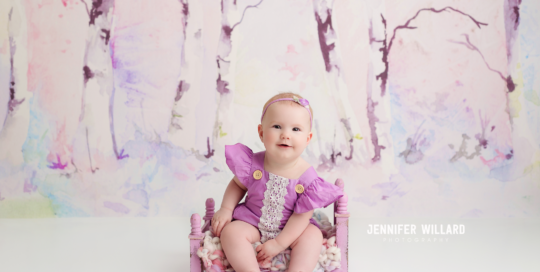 baby girl in purple romper on purple bed with watercolour tree backdrop